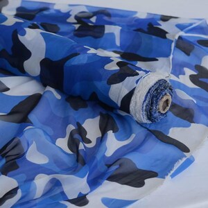 Puede incluir: Un rollo de tela semitransparente con un patrón de camuflaje en tonos azul real, azul claro, blanco y negro. El material está enrollado firmemente alrededor de un núcleo de cartón, con tela suelta drapeada debajo de forma fluida.