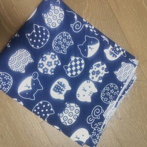 Könnte beinhalten: Ein gefaltetes Stück marineblauen Stoffes mit einem sich wiederholenden Muster aus weißen Katzenköpfen. Die Katzenköpfe haben verschiedene Designs, darunter Punkte, Blumenmuster und geometrische Formen. Der Stoff ist wahrscheinlich Baumwolle oder ein ähnliches Material.