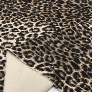 May include: Close-up of a fabric with a leopard print pattern. The fabric features a tan background with dark brown and black spots. The reverse side of the fabric is a solid beige color. The fabric is folded over, revealing the underside.