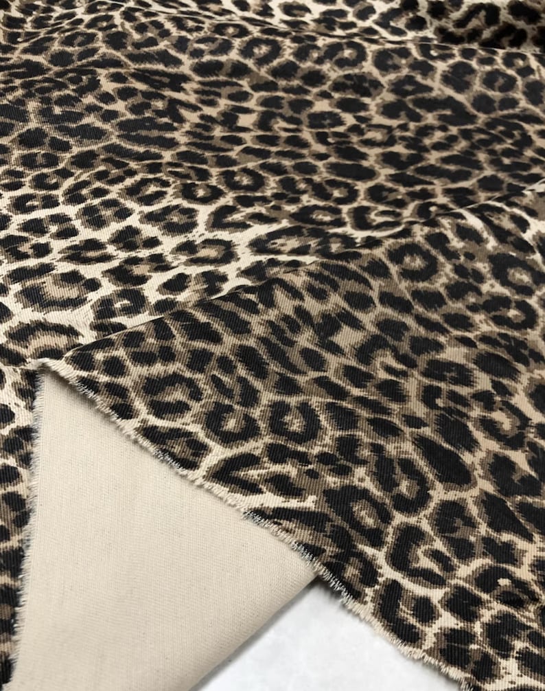 May include: Close-up of a fabric with a leopard print pattern. The fabric features a tan background with dark brown and black spots. The reverse side of the fabric is a solid beige color. The fabric is folded over, revealing the underside.