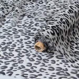 Puede incluir: Un rollo de tela semitransparente con un patrón repetitivo de estampado de leopardo en negro y gris sobre un fondo blanco. El tejido está enrollado en un tubo de cartón, mostrando pliegues y caídas que resaltan la calidad translúcida del material.