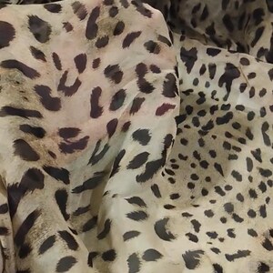 Puede incluir: Una bufanda ligera y semitransparente con un estampado de leopardo clásico. La tela muestra un diseño repetitivo de manchas irregulares de color marrón oscuro sobre un fondo beige claro, creando una apariencia translúcida y fluida.