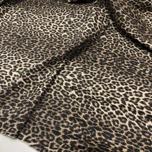 May include: Close-up of a fabric with a leopard print pattern. The fabric features a tan background with dark brown and black spots. The material is draped, showing folds and texture.