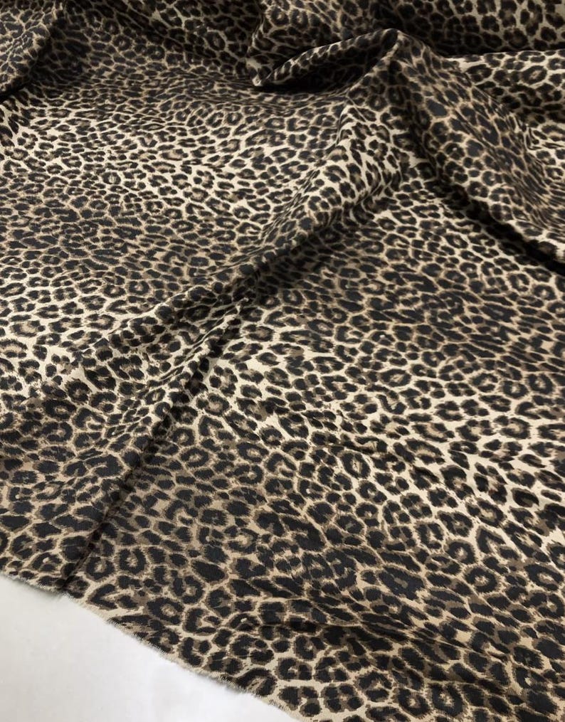 May include: Close-up of a fabric with a leopard print pattern. The fabric features a tan background with dark brown and black spots. The material is draped, showing folds and texture.