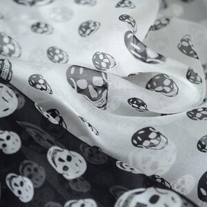 Könnte beinhalten: Ein leichter Schal mit einem sich wiederholenden Muster aus schwarzen Totenköpfen auf weißem Grund und weißen Totenköpfen auf schwarzem Grund. Der Stoff hat eine halbtransparente, glatte Textur mit weichen Falten, die Tiefe und Bewegung erzeugen.