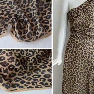 Puede incluir: Una tela transparente con un estampado de leopardo en tonos beige y marrón. El material se muestra drapeado en pliegues y sobre un maniquí como una prenda de un solo hombro. La tela tiene un borde acabado y una calidad semitransparente.