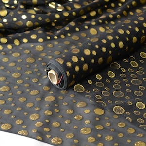 May include: A roll of sheer black fabric featuring a repeating pattern of metallic gold circles of varying sizes. The fabric is draped to show the translucent quality of the material and the textured, shimmering appearance of the gold foil dots.