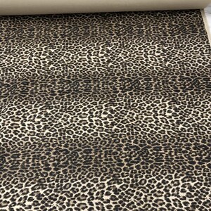 May include: A roll of fabric featuring a leopard print pattern. The fabric has a beige background with dark brown and black spots. The roll is partially unrolled, revealing the fabric's texture and design.