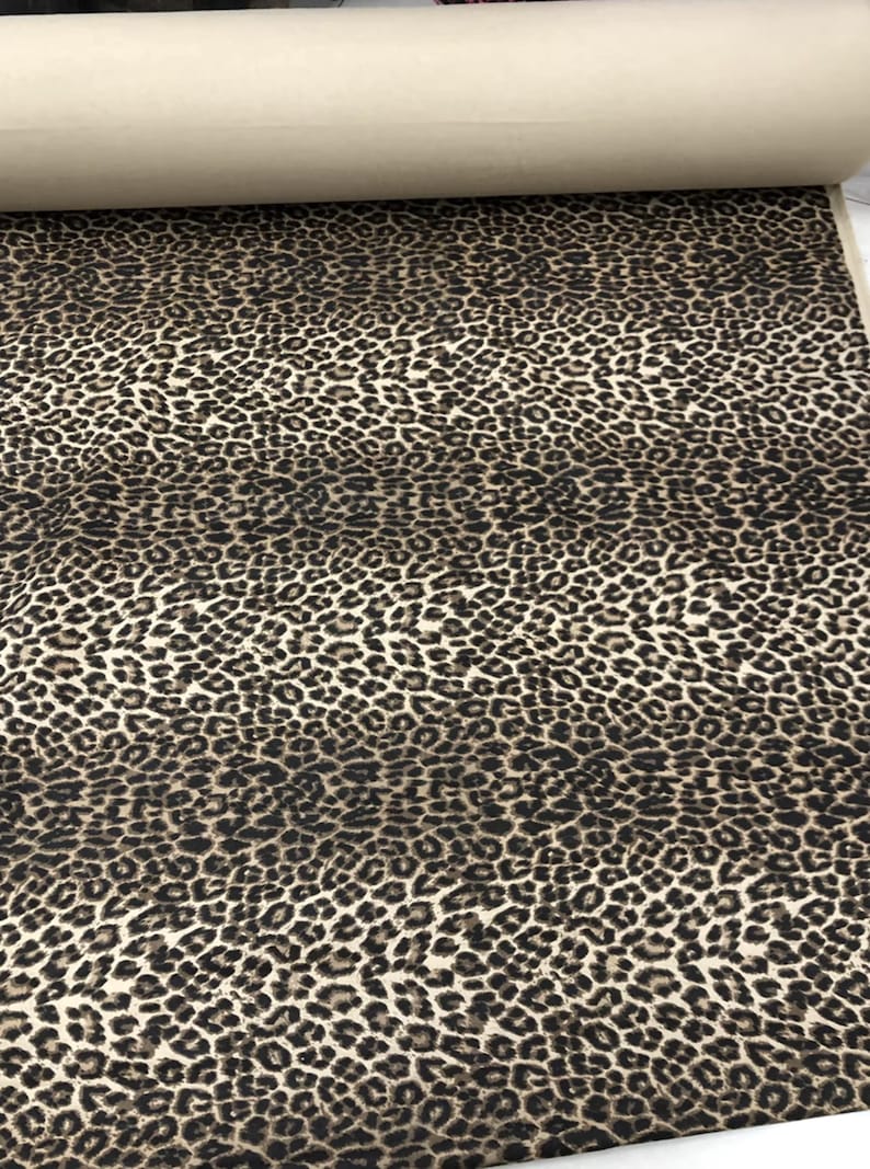 May include: A roll of fabric featuring a leopard print pattern. The fabric has a beige background with dark brown and black spots. The roll is partially unrolled, revealing the fabric's texture and design.