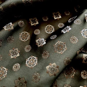 May include: Close-up of a dark green fabric with a repeating floral pattern. The flowers are in shades of brown, beige, and white, creating a subtle contrast against the deep green background. The fabric is folded, showing the texture and sheen.