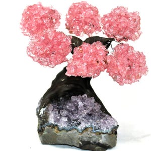 May include: A decorative crystal tree with pink crystal blossoms and a dark gray trunk. The base is a gray rock formation with purple crystals. The tree is a unique home decor item.