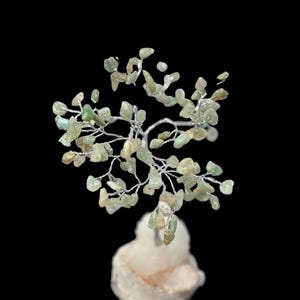Puede incluir: Escultura decorativa de árbol con estructura de alambre plateado y pequeñas hojas de piedras preciosas verde claro. El árbol se asienta sobre una base de piedra rugosa blanquecina sobre un fondo negro. Las piedras tienen forma natural.