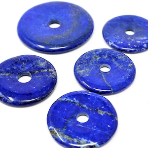 Puede incluir: Cinco piedras pulidas en forma de rosquilla de lapislázuli azul profundo, con un agujero central. Las piedras tienen vetas naturales doradas y variaciones de color, sobre un fondo blanco. Estas piedras preciosas son adecuadas para la fabricación de joyas.