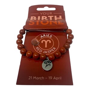 Może przedstawiać: Bransoletka z koralików z czerwonego jaspisu ze srebrnym charms z symbolem zodiaku Barana. Bransoletka jest prezentowana na czerwonej karcie z napisem "Your Birth Stone" i "Aries Red Jasper 21 March - 19 April."