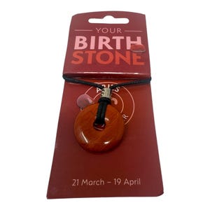 Może przedstawiać: Naszyjnik z wisiorkiem z czerwono-brązowego kamienia z czarnym sznurkiem, prezentowany na czerwonej karcie. Karta zawiera napis "YOUR BIRTH STONE" i "ARIES" z datami "21 marca - 19 kwietnia". Wisiorek ma okrągły kształt z otworem pośrodku.