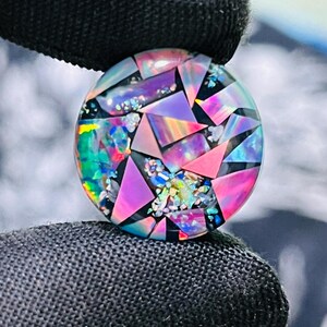 May include: A round, iridescent gemstone with a mosaic design. The gemstone features a mix of pink, purple, blue, and green triangular and irregular shapes set against a black background. The gem is held by a black gloved hand.
