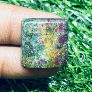 Könnte beinhalten: Ein quadratisch geschliffener Edelstein mit gesprenkeltem Aussehen. Der Stein weist eine Mischung aus grünen, gelben und roten Farbtönen auf und hat eine leicht raue Textur. Der Edelstein wird in einer Hand gehalten, mit einem Hintergrund aus grünem Gras.