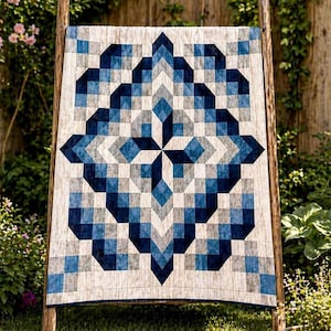 May include: A geometric quilt with a symmetrical design in shades of blue, gray, and white is displayed on a wooden rack outdoors. The quilt features a central starburst pattern and a border of alternating squares. The background includes greenery and a wooden fence.