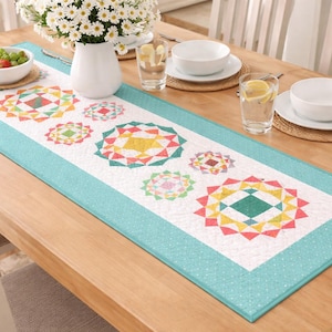 Op de afbeelding: Een rechthoekige tafelloper met een wit midden met kleurrijke geometrische ontwerpen in rood, geel, groen en turquoise. De loper heeft een turquoise rand met een stippenpatroon en ligt op een houten tafel.