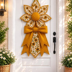 May include: A quilted fabric door hanging shaped like a large sunflower with a central mustard yellow disc, petals featuring a sunflower print on a cream background, and a large mustard yellow quilted bow with long, pointed fabric tails.