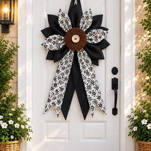 Può includere: Una ghirlanda decorativa per porta con un grande design a fiore con petali neri solidi alternati a petali bianchi con un motivo floreale nero. Il centro è una bobina di corda marrone con un grande bottone beige. Due lunghi nastri pendono.