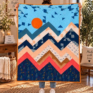 May include: A rectangular quilt featuring a mountain landscape design with a bright orange sun against a light blue sky. The mountains are composed of layered chevron patterns in shades of dark blue, peach, tan, and coral, with a forest print base.