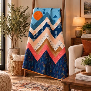 Könnte beinhalten: Eine Steppdecke mit Berglandschaftsmotiv, bestehend aus einer leuchtend orangefarbenen Sonne, blauem Himmel und zickzackförmigen Gipfeln in Pfirsich-, Beige- und Weißtönen. Der untere Bereich zeigt einen dunkelblauen Waldaufdruck mit kleinen Hirschen.