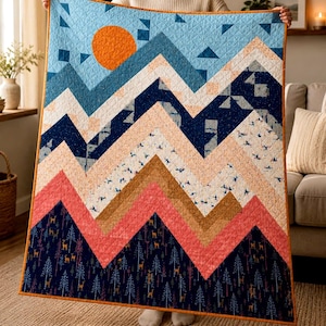 May include: A colorful quilt with a mountain range design. The quilt features a blue sky with an orange sun, and layers of blue, peach, and brown fabric. The bottom layer has a navy blue forest pattern with deer and trees. The quilt is trimmed with an orange border.