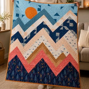 Puede incluir: Una colcha rectangular con un diseño de paisaje montañoso y un sol naranja brillante en un cielo azul claro. Las montañas están compuestas por patrones de chevrón en tonos azul oscuro, crema, bronceado y coral, con un estampado de bosque.