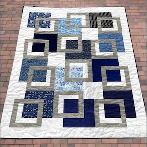 May include: A handmade quilt featuring a geometric design with squares and rectangles in shades of blue, white, and gray. The quilt has a white background with quilted patterns and a black border.