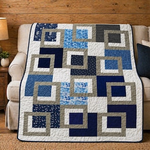 May include: A handmade quilt with a geometric design in shades of blue, gray, and white. The quilt features a square-in-square pattern, with various blue floral and dotted fabrics. The quilt is draped over a beige sofa.