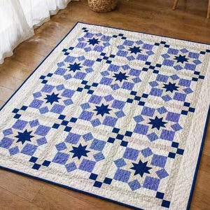 May include: A rectangular quilt featuring a repeating pattern of eight-pointed stars in shades of dark blue and light blue on a white background. The design includes small blue square accents between the stars and a solid dark blue border edge.