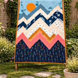 May include: A colorful quilt with a mountain landscape design. The quilt features a blue sky with an orange sun, layered mountain peaks in shades of blue, pink, and tan, and a forest pattern at the bottom. The quilt is displayed on a wooden stand.