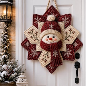 May include: A quilted star-shaped wall hanging featuring a central snowman face wearing a dark red knit hat with a white pom-pom and a matching fringed scarf. The star has alternating dark red and cream panels with embroidered snowflake patterns.
