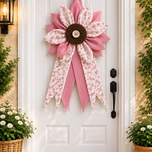 May include: A fabric door hanging shaped like a large flower with alternating petals of solid pink and pink floral print. The centre is a dark brown circular disc with a light beige button. Two long fabric tails hang down from the flower base.