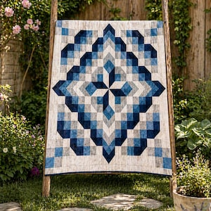 May include: A geometric quilt in shades of blue, gray, and white is displayed on a wooden rack outdoors. The quilt features a symmetrical pattern of squares and diamond shapes, creating a star-like design. The quilt is displayed on a wooden rack in a garden setting.