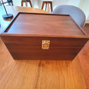May include: A rectangular wooden box with a hinged lid and a gold-colored clasp. The box is made of dark brown wood and sits on a wooden surface. The box appears to be empty.