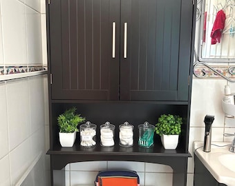 May include: A dark brown over-the-toilet storage cabinet with two doors and a lower shelf. The shelf holds small jars, plants, and a cosmetic bag. The cabinet has silver handles and is in a bathroom setting.