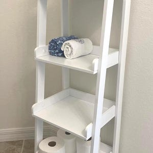 May include: A white, four-tiered wooden shelf unit with a ladder-style design. The shelves hold decorative plants, rolled towels, and toilet paper. The unit is designed for bathroom storage and organization.