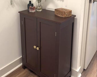 May include: A dark brown wooden cabinet with two doors and gold-colored knobs. The cabinet is rectangular and has a flat top. Decorative items, including a small box and bottles, sit on top of the cabinet. The cabinet is in a room with light-colored walls and a wooden floor.
