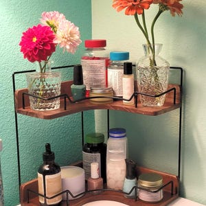 May include: A two-tiered corner shelf made of dark wood and black metal, holding various items. The top shelf displays a vase of pink and white flowers, nail polish, and bottles. The bottom shelf has a candle, bottles, and jars. The background is a light teal wall.