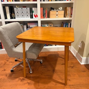 May include: A light brown wooden desk with four legs. A gray upholstered office chair with a chrome base is positioned behind the desk. The desk is in front of a white bookshelf filled with books and decorative items.