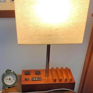 May include: A wooden table lamp with a rectangular beige lampshade. The lamp has a wooden base with electrical outlets and USB ports. A small, green, antique-style clock sits on the table.
