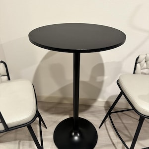 May include: A black round bistro table with a single central support and a circular base. Two chairs with black metal frames and white cushioned seats flank the table. The table is approximately 30 inches in height.