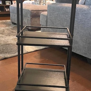 May include: A black metal serving cart with three shelves. The cart has a rectangular frame and is mounted on four small wheels. The shelves are made of dark wood. The cart is designed for use in a living room or dining area.