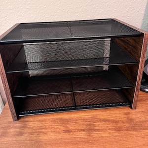 May include: A three-tiered desktop organizer with a dark wood-grain finish and black mesh shelves. The organizer is designed to hold papers, files, or other office supplies. The sides and top are made of wood, while the shelves are made of metal mesh.