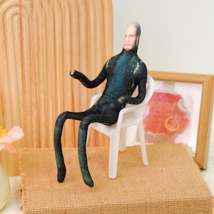 Puede incluir: Muñeco de tela de una persona con un mono azul oscuro y negro, sentado en una pequeña silla de plástico blanca. El muñeco tiene la cara pálida y el pelo gris. El muñeco está sobre una pequeña plataforma cuadrada cubierta de arpillera.