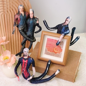 May include: A collection of handmade fabric dolls with detailed facial features and outfits. The dolls are posed in various positions, some interacting with a small framed picture. The color palette includes red, blue, black, and white.