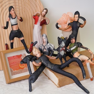 Pode incluir: Uma coleção de bonecas de tecido feitas à mão, com personagens de um videogame. As bonecas têm traços faciais detalhados e estão vestidas com várias roupas. As bonecas estão dispostas em uma superfície de madeira com uma foto emoldurada e um livro.