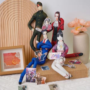 May include: A collection of handmade fabric dolls and character cutouts. The dolls are dressed in various outfits, including a blue suit with a red tie, a red coat, and a green military-style uniform. The cutouts feature various characters.